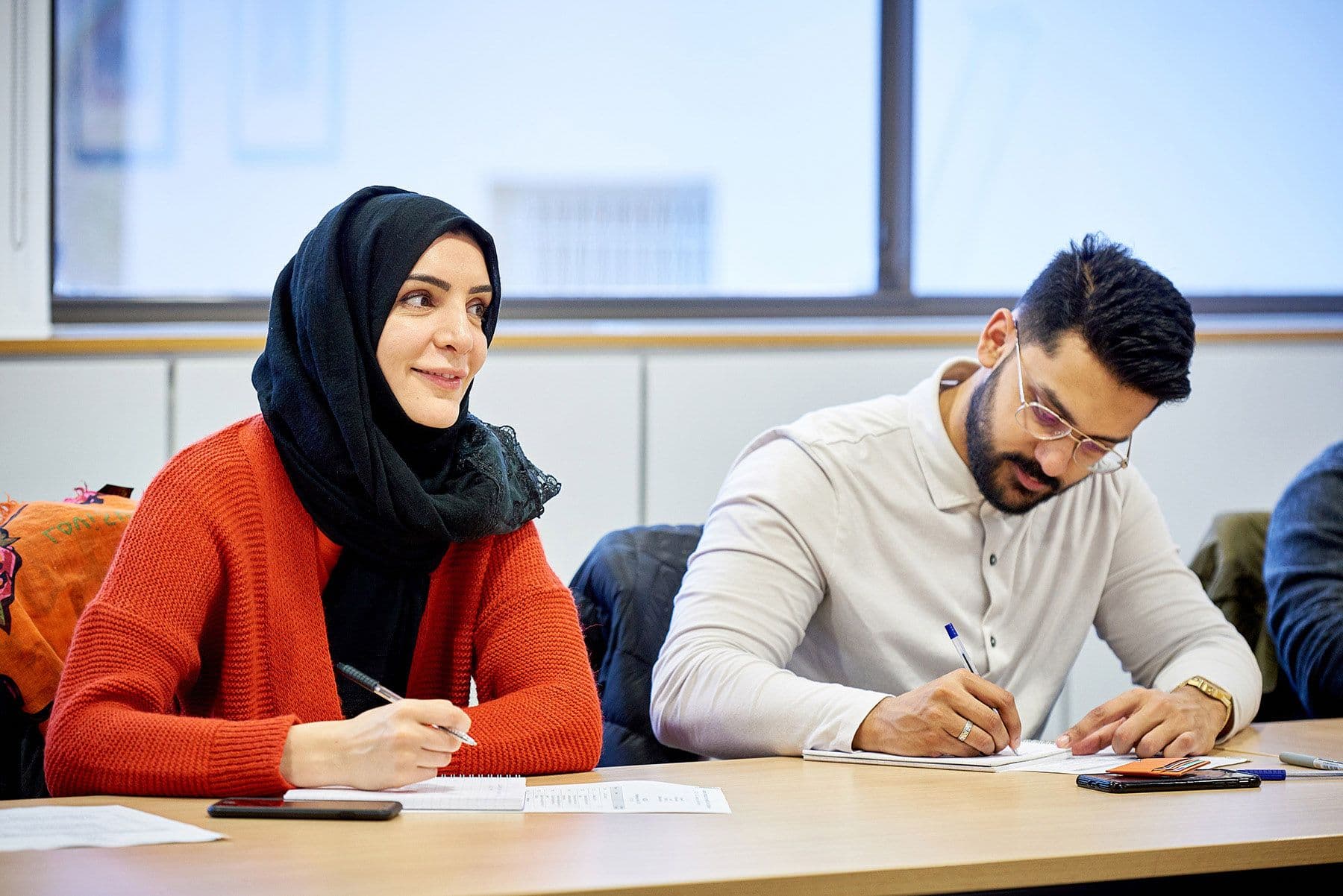 A woman wearing a hijab and a man writing notes together in a meeting.