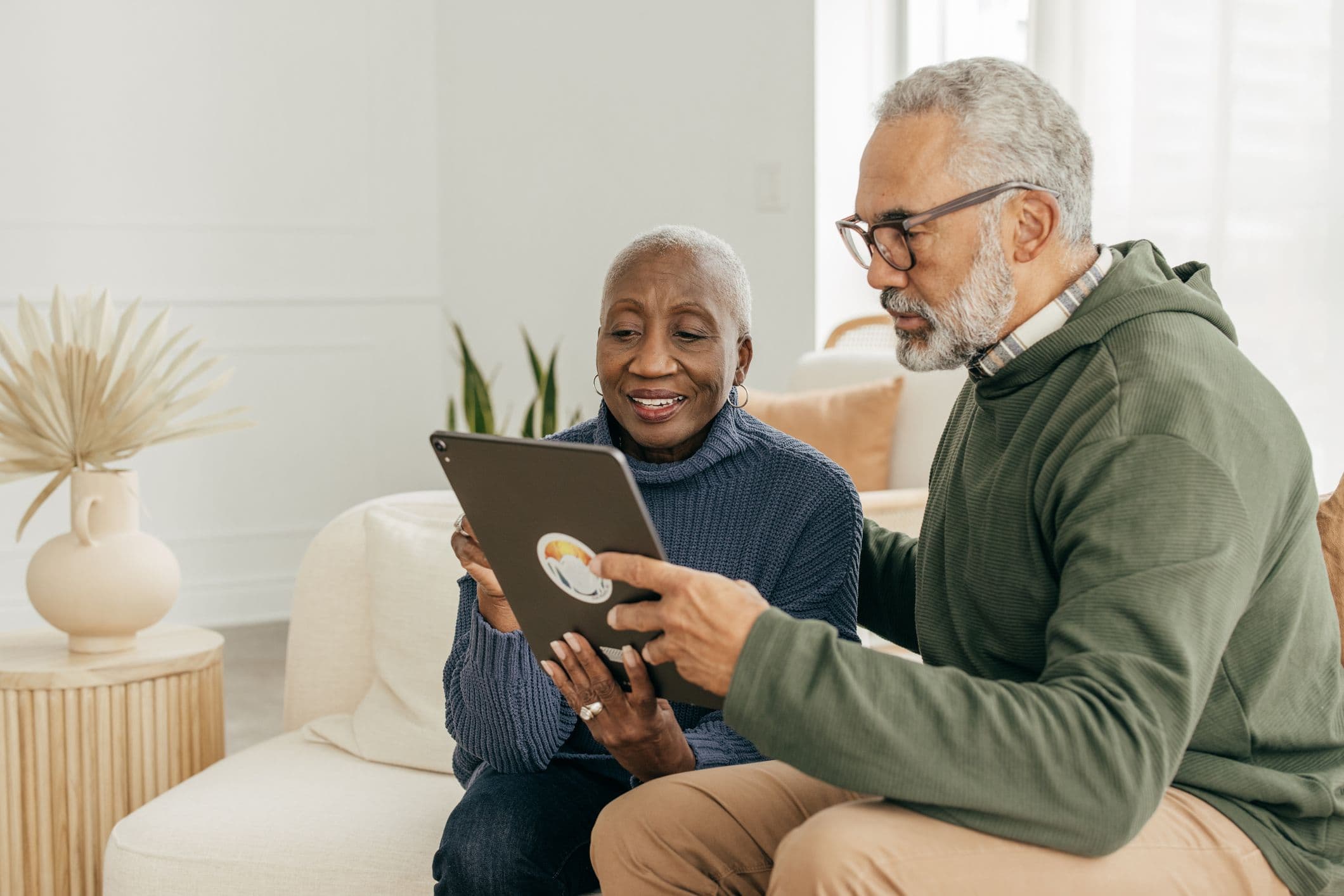 Two older adults sitting on a sofa, smiling and looking at a tablet together in a cozy, well-lit room with plants and decorative elements.