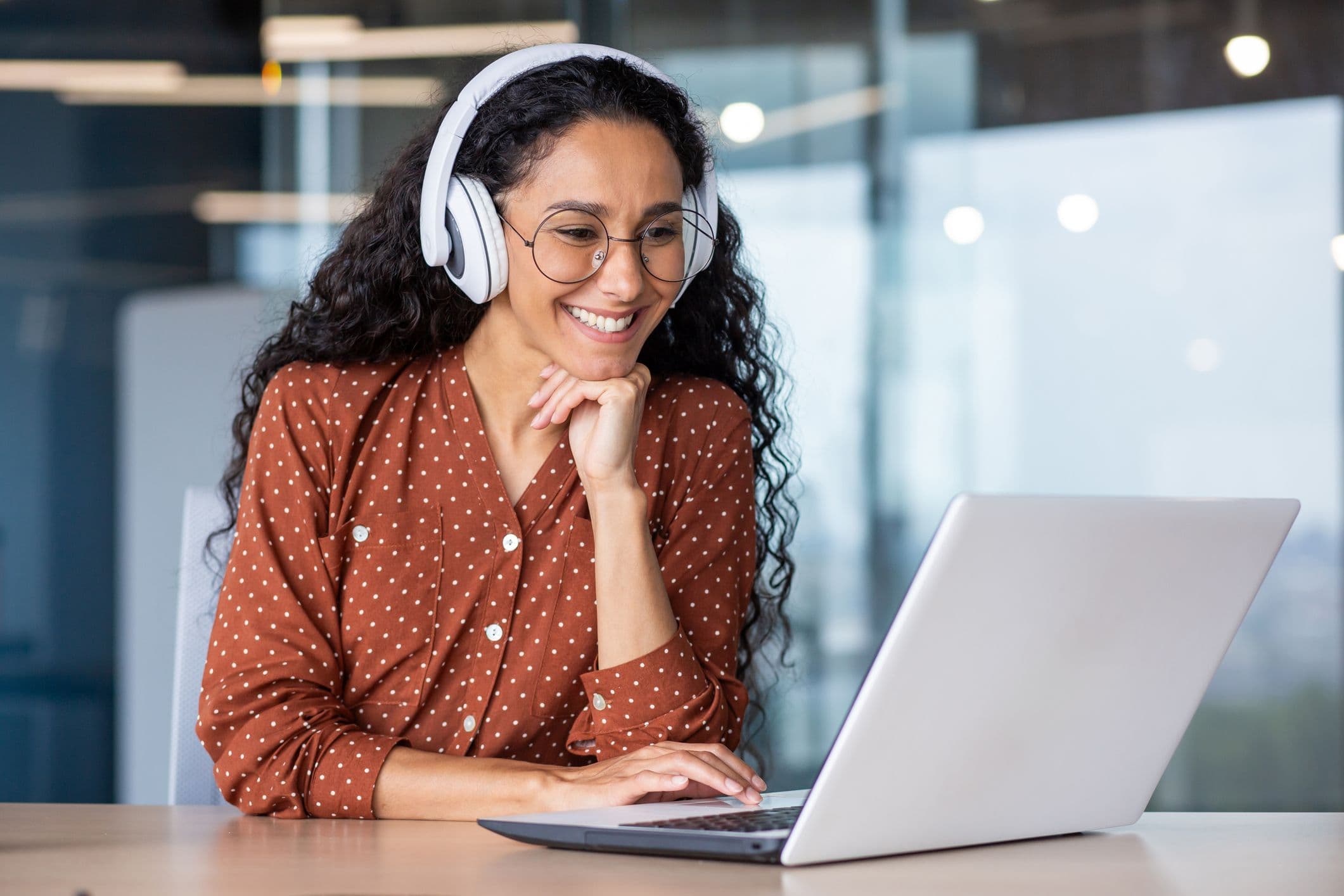 Smiling woman with headphones and glasses uses a laptop in an office, wearing a brown polka dot shirt.