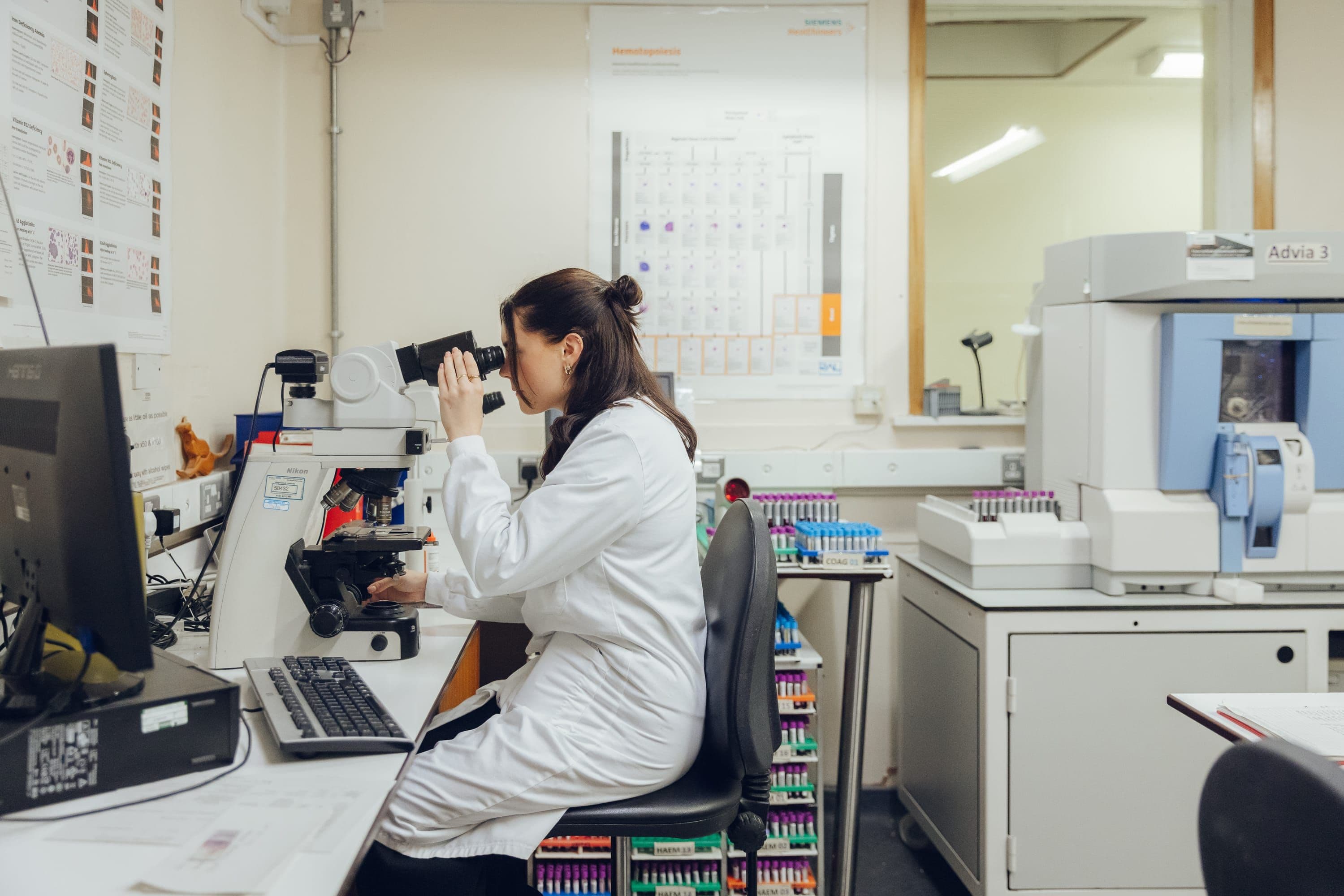 A scientist in a lab coat observes samples through a microscope in a laboratory filled with various equipment and supplies.