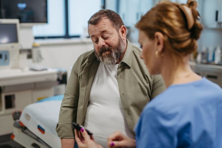 A man in a white shirt and green jacket sits on an examination table while a medical professional in blue scrubs shows him something on a device.
