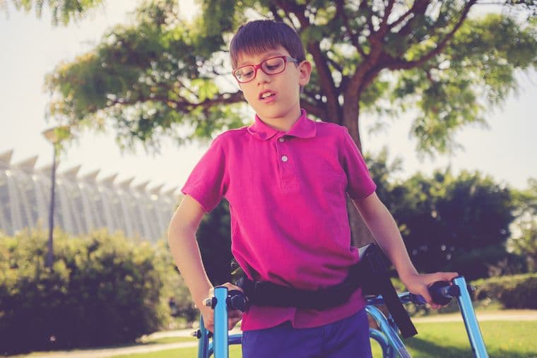 A young boy with glasses uses a walker in a sunny park, wearing a bright pink shirt, with trees and a modern building in the background.
